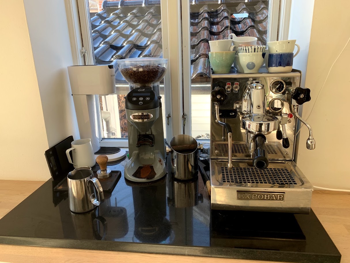 Espobar espresso machine, grinder, coffee beans, and milk pitchers arranged on a countertop by a window.
